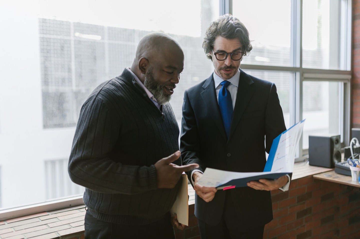 Two men standing by a window reviewing the papers in an open blue folder